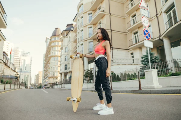 スタイリッシュなストリートウェア姿のライダーガールが、ロングボードを手にしたカメラでポーズをとり、街中で真剣な顔をしたカメラを見る。スケートボードを持つ少女のストリートポートレート。 — ストック写真