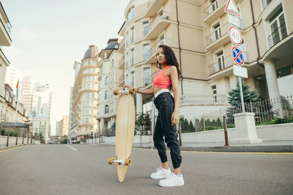 Chica jinete de moda en ropa de calle con estilo posa en la cámara con longboard en la mano, mira a la cámara con cara seria en el paisaje urbano. Retrato callejero de una chica con monopatina.Longboard —  Fotos de Stock