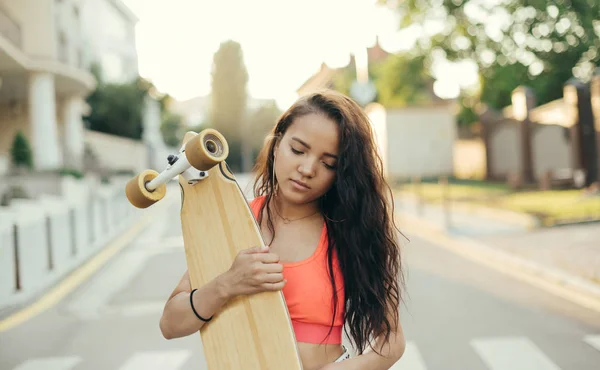 Retrato de close-up de menina atraente andando com longboard em mãos na rua contra o fundo do pôr do sol, olhando para baixo. Uma rapariga de rua com um quadro negro nas mãos. Estilo de vida photo.Extreme esportes — Fotografia de Stock