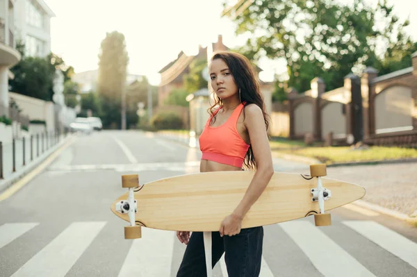 Muchacha hispana atractiva caminando con un longboard en sus manos en las calles de una ciudad nocturna, mirando en cámara, usando ropa de calle con estilo. Retrato de una chica con un monopatín —  Fotos de Stock