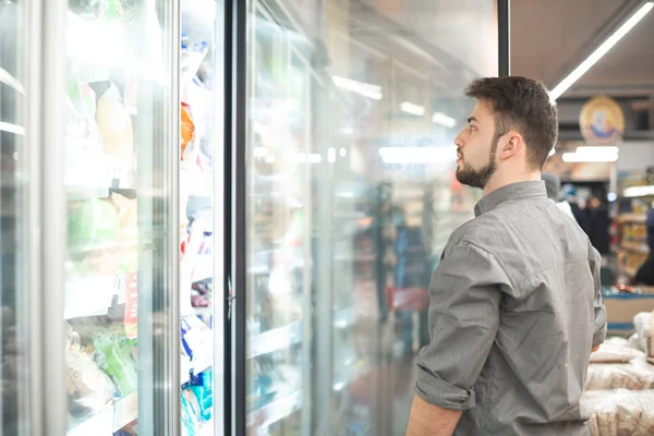 Um homem escolhe alimentos congelados de prateleiras em um refrigerador em um s — Fotografia de Stock