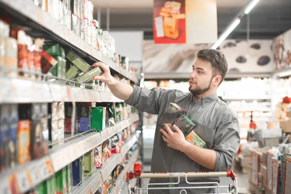 Buyer buys a discount product at a supermarket. Portrait of a man wearing a shirt, takes packets of tea from supermarket shelves.Man with a shopping cart takes a lot of tea packs in the grocery store — Stock Photo, Image