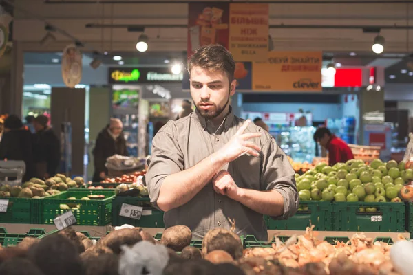 El hombre con barba se para en el departamento de verduras de un supermercado con guantes en las manos y elige verduras. El barbudo compra verduras y un supermercado. Compras en una tienda de comestibles — Foto de Stock