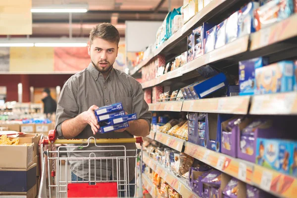 Hombre con un paquete de galletas en las manos y carrito de la compra en un — Foto de Stock