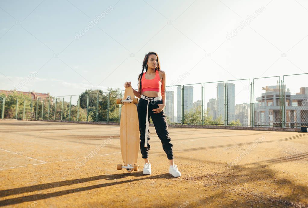 Attractive girl in trendy street clothes stands with a longboard in hand on the playground, posing at the camera,looking away. Hispanic girl model with skateboard in hands stands on old tennis court.