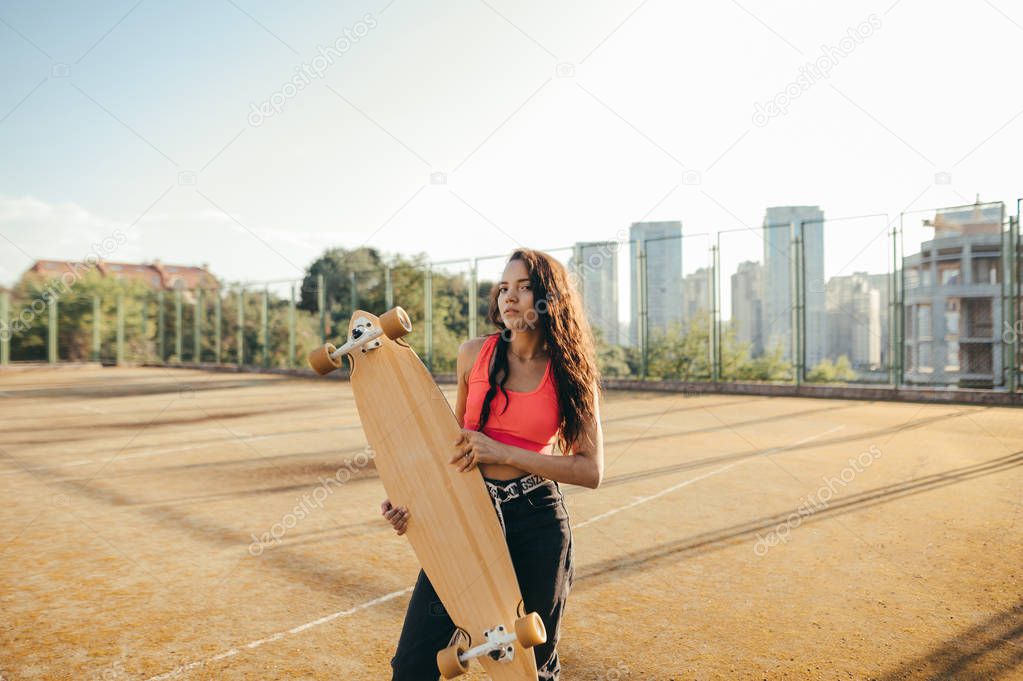 Street portrait of a mulatto stands on the playground,holds a longboard in hands, looks into the camera, wears stylish casual clothes. Curly hispanic girl model posing with a longboard in her hands.