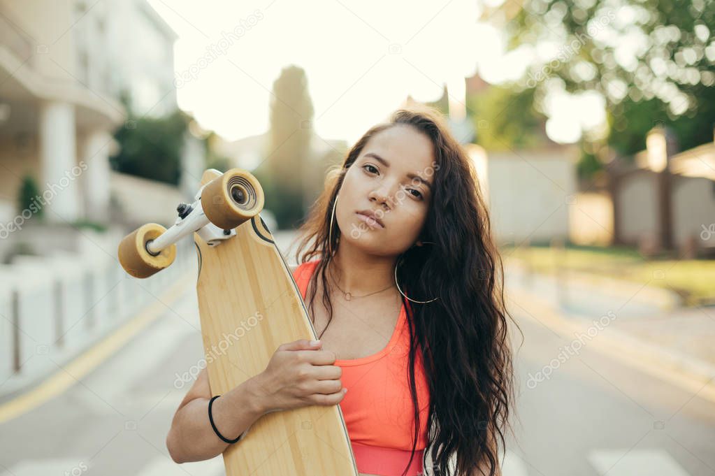 Street portrait of a mulatto girl holding a longboard in her hands, looking in camera, walking with a board in the evening. Girl rider with skateboard in hand stands on the street