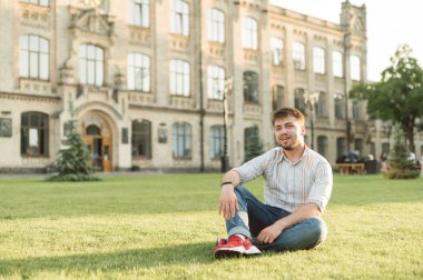 Üniversite binasının arka bahçesinde çimüzerinde oturan sakallı mutlu bir öğrenci, gülümseyerek ve uzağa bakıyor, gömlek giyiyor. Kampüs kampüsünde dinlenen yetişkin bir erkek öğrencinin portresi.