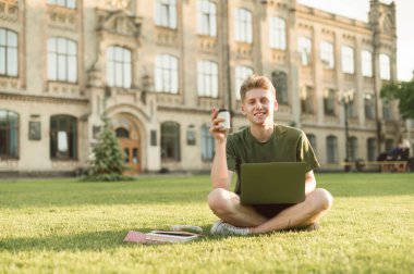 Gülümseyen genç adam kucağında bir dizüstü bilgisayar ile bir üniversite kampüsünde bir çim oturur, elinde bir fincan kahve tutar, kameraya bakar. Molada dinlenen öğrencinin portresi