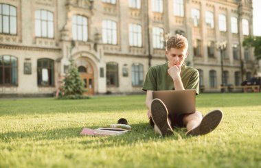 Düşünceli bir adam bir dizüstü bilgisayar ile üniversitenin arka bahçesinde çim üzerinde oturan, düşünceli ekrana bakarak ve eğitim. Kampüste dizüstü bilgisayarla çalışan genç bir öğrencinin portresi.