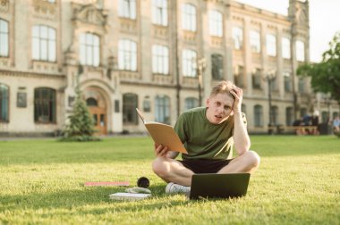 Şaşkın genç erkek öğrenci dizüstü bilgisayar ve kitaplar ile üniversite çim oturan çalışmaya çalışıyor. Ev ödevi, deneme yazma ve tez ile ilgili sorunlar olan sinirli üniversite öğrencisi.