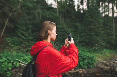 Yürüyüşçü kız dağ nehrinin yanındaki ormanda duruyor ve kırmızı yağmurluk giyip akıllı telefondan manzara fotoğrafı çekiyor. Çekici kadın, akıllı telefon kamerasıyla dağlardaki doğal manzaraların fotoğraflarını çekiyor.