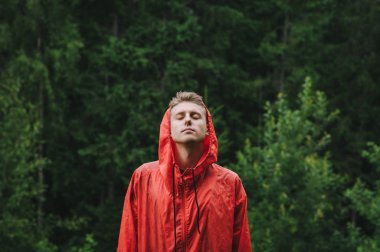 Kırmızı yağmurluk giyen yakışıklı genç adam yağmurun altında gözleri kapalı ve yağmurun tadını çıkararak dışarıda duruyor. Yürüyüş yapan genç bir erkek yürüyüşçünün portresi. Yeşil bir arka planda yağmurda duruyor..