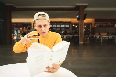 Şapkalı ve sarı kazaklı zeki bir adam elinde bir sandviçle yemek masasında oturur ve kitap okur. Öğrenci bir kafede kitap okur ve fast food yer. Öğrencinin hiç boş vakti yok..