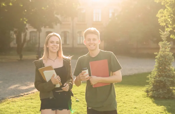 Dos estudiantes emocionados con exámenes aprobados cerca de la universidad sosteniendo los cuadernos y llevar la taza de café. Un par de estudiantes en el parque cerca de la universidad sonriendo y sintiéndose descuidados . — Foto de Stock