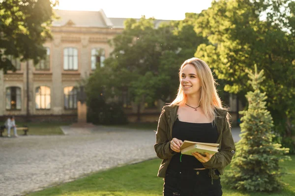 Krásná studentka s širokým úsměvem drží zápisníky a dívá se stranou do univerzitní zahrady. Usměvavá studentská dívka s knihami a učebnicích v parku nedaleko univerzity. — Stock fotografie