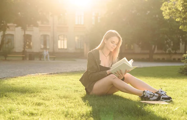 Hermosa joven estudiante sentada en el césped en el parque universitario leyendo el libro. Estudiante alegre sentada en la hierba en el jardín de la universidad con un libro. Concepto de educación, espacio de copia . — Foto de Stock