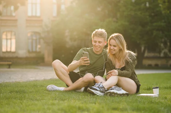 Happy caucasian college students using smartphone on campus lawn. Cheerful couple of students watching video on the phone, using social media on the cell phone in the university park.