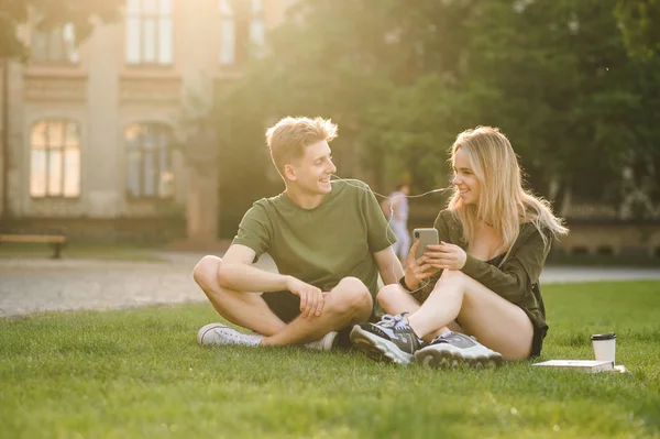 Jovem casal de estudantes universitários relaxar e ouvir música do smartphone com fones de ouvido na grama no parque. Dois estudantes alegres sentados no gramado perto da universidade desfrutando de música em fones de ouvido . — Fotografia de Stock
