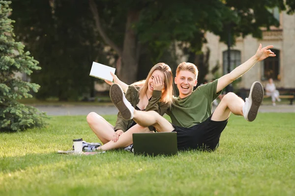 Два счастливых веселых студента валяют дурака, сидя на лужайке колледжа с книгами, ноутбуком и забирая кофе. Пара студентов университета развлекаются на траве возле кампуса с учебниками и ноутбуками — стоковое фото