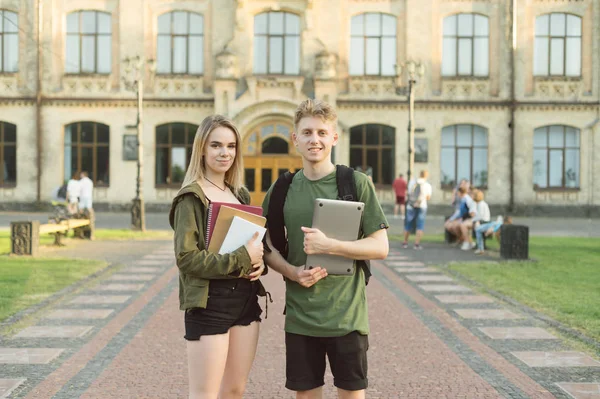 นักศึกษามหาวิทยาลัยสองคนที่มีความสุขที่น่าสนใจพูดคุยกันที่สวนสาธารณะวิทยาลัยถือหนังสือคัดลอกและแล็ปท็อป คู่นักเรียนที่มีความสุขที่ยิ้ม ยืนอยู่ใกล้มหาวิทยาลัย พร้อมหนังสือเรียน หนังสือ และคอมพิวเตอร์ . — ภาพถ่ายสต็อก