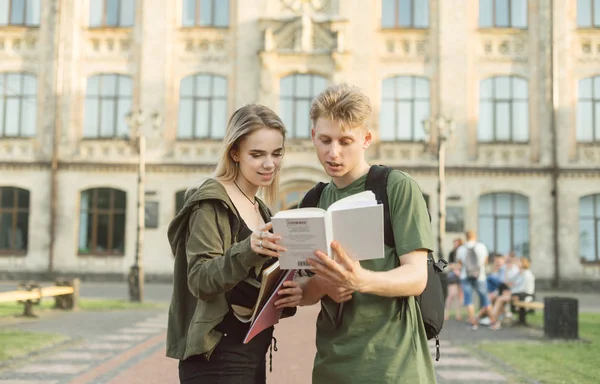 Young attractive cute students couple standing in front of the university reading an interesting book. Two concentrated amazed students reading the book near the college preparing for classes.