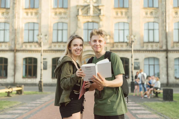 Щасливі веселі двоє студентів в університетському містечку тримають книгу, дивлячись на камеру. Приваблива позитивна пара студентів, що стоїть перед будівлею коледжу з підручником . — стокове фото