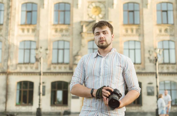 Portré egy fiatal csinos szakállas férfi csíkos ing Holding professzionális kamera, lövészet a szabadban, stílusosan öltözött, fotós, közelről, brutális, utcai fotó, architektúra háttér. — Stock Fotó