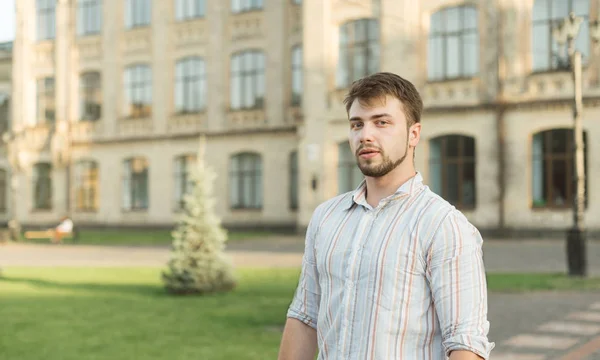 Masculine bearded male college student standing in front of the campus building in the park. Attractive serious student man with beard looking at camera near the university in the garden.