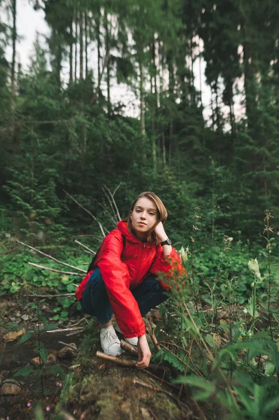 빨간 비옷을 입은 아름다운 소녀 가산 위 로그 위에 앉아, 진지 한 얼굴로 카메라를 들여다보고 있다. 산속의 숲 속에서 앉아서 쉬고 있는 관광객의 사진. — 스톡 사진