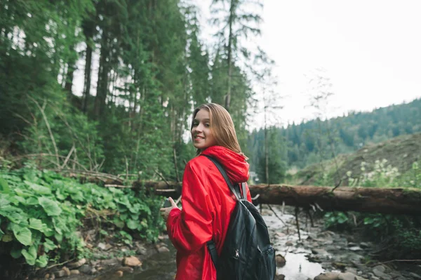 Retrato de chica senderista sonriente caminando, mirando a la cámara y sonriendo en el fondo del arroyo y las montañas, vistiendo impermeable rojo. Descanso activo, caminar por las montañas, turismo de montaña . — Foto de Stock