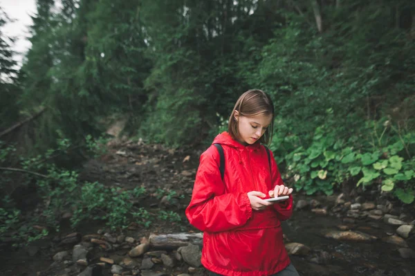 빨간 비옷을 입은 아름다운 소녀는 산악 지대와 숲의 배경에서 스마트폰을 사용 합니다. 지도를 보고 스마트폰을 이용하고 산악 지역을 하이킹하는 여성 이 있습니다. 야생의 기술 — 스톡 사진