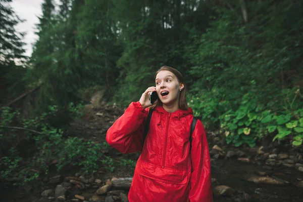 빨간 자켓을 입은 표현력 있는 여행객 이 산등성 이 배경에 서서, 전화로 말하고 놀란 얼굴로 놀란 표정을 짓고 있다. 놀라운 관광객들이 아름다운 야생 동물 숲이라고 부르다. — 스톡 사진