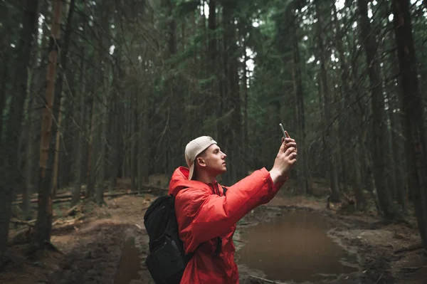 Jovem de casaco vermelho fica na floresta durante uma caminhada e tira uma foto em uma câmera de smartphones. Retrato de homem caminhante na floresta com telefone na mão no fundo da trilha da montanha com poça. . — Fotografia de Stock