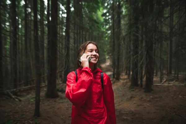 행복 한 관광객 소녀가 등산중 전화 통화를 하고, 숲 속에서 서, 빨간 자켓을 입고 배낭을 찬 채, 시선을 돌리며 미소짓고 있습니다. 긍정적 인 관광 여성 하이킹 사진, 전화 통화. — 스톡 사진