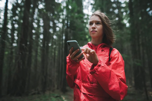 Chica concentrada en una chaqueta roja se para por la noche en el bosque con un teléfono inteligente en las manos y mira hacia otro lado con ansiedad. Chica turística utiliza teléfono inteligente en una caminata, mira los mapas y la navegación . — Foto de Stock