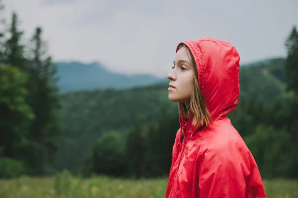 Портрет крупним планом в профілі привабливої дівчини в червоному плащі стоїть на тлі гір і хвойних лісів в похмуру дощову погоду, дивлячись з серйозним обличчям . — стокове фото