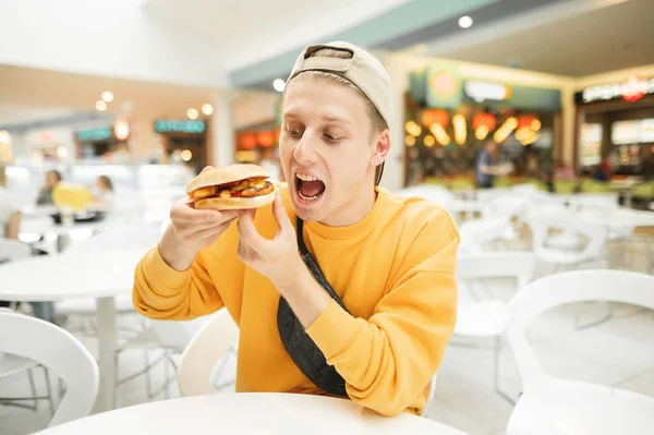 밝은 색 옷을 입은 쾌활 한 청년 이 시장의 카페에서 식욕을 돋우는 샌드위치를 입고 배고픈 표정으로 패스트푸드를 찾는다. 배고픈 학생 이 햄버거를 먹다. — 스톡 사진