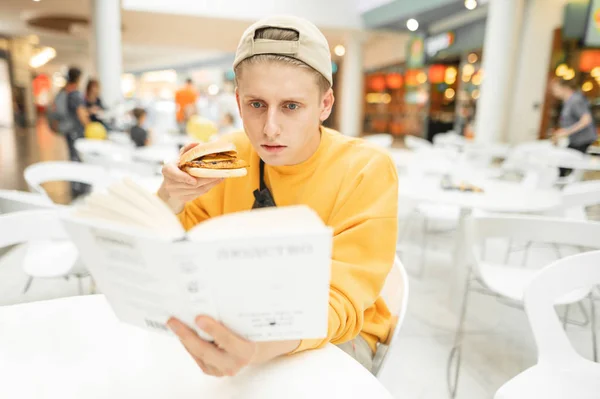 Ritratto di giovane concentrato in abbigliamento casual e berretti, leggere un libro e mangiare un panino. Un ragazzo con un hamburger in mano legge un libro in un fast-food — Foto Stock