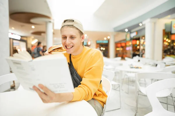 Ευτυχισμένο αγόρι με μπιφτέκι στο χέρι κάθεται σε ένα εστιατόριο στο λευκό τραπέζι, κοιτάζει το βιβλίο και χαμογελαστά.Θετική φοιτητής σπουδάζει σε ένα καφέ και δείπνο με fast food, φορώντας ένα φωτεινό casual ρούχα. — Φωτογραφία Αρχείου