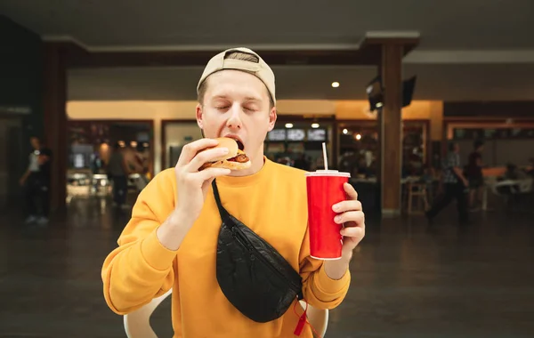Hungry guy in a street clothing and cap sits in a cafe and eats a sandwich holding a glass of cola in his hand.Young man biting a burger with closed eyes against the backdrop of a fast food restaurant — Stock Photo, Image