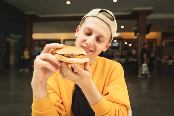 Retrato cercano de un chico satisfecho con una hamburguesa en la mano, hambriento mirando un sándwich y quiere comerlo, fondo de un restaurante de comida rápida en un centro comercial. — Foto de Stock