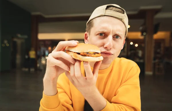 Foto divertida de un joven alegre con gorra y un suéter amarillo sentado en un restaurante de comida rápida, mira a la cámara y hace una cara divertida. Retrato cercano de un estudiante con un sándwich en la mano . — Foto de Stock