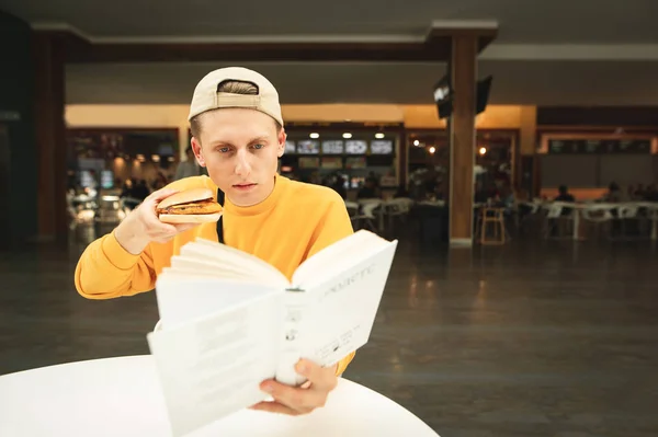 Un tipo listo con una gorra y un suéter amarillo se sienta a la mesa en un patio de comidas con un sándwich en la mano y lee un libro.El estudiante lee un libro y come comida rápida en un café. El estudiante no tiene tiempo libre . — Foto de Stock