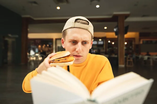 Close portrait of a good student reads a book and eats a burger at a fast food restaurant. Guy prepares for an exam in a cafe, eats a snack and looks at the book. Fast food and student