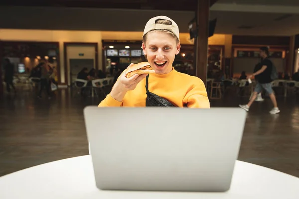 Счастливый мальчик с бургером в руке, глядя на экран ноутбука и улыбаясь, сидит за столом на фоне ресторана быстрого питания. Студент ест сэндвич и пользуется ноутбуком . — стоковое фото