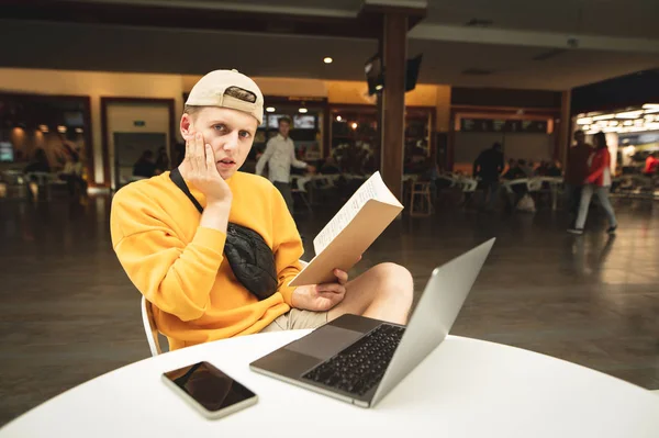 Retrato de un estudiante frustrado sentado en un café con un portátil y un cuaderno en la mano, mirando a la cámara con una mirada triste al fondo de los restaurantes de comida rápida . — Foto de Stock