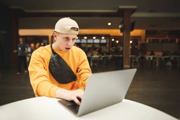 Эмоциональный молодой человек фрилансер в повседневной одежде работает в торговом центре на фоне ресторанов быстрого питания, в кепке и поясной сумке, смотрит на экран ноутбука с изумлением . — стоковое фото