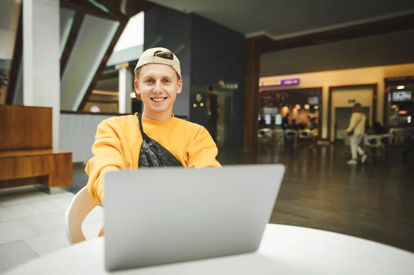 Забавный парень в стильной уличной одежде работает над ноутбуком в торговом центре, смотрит в камеру и улыбается. Счастливый фрилансер работает на ноутбуке в торговом центре — стоковое фото