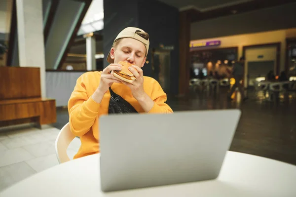 Jovem em um vestido casual sentado em uma mesa em um restaurante de fast-food, morde um hambúrguer com os olhos fechados e trabalha em um laptop. Almoço enquanto trabalhava na Internet . — Fotografia de Stock